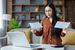 © Liubomir - Asian woman analyzing papers at home, wearing glasses, using laptop. Focusing on important details, multitasking in living room. Concept of concentration, home office, paperwork management.