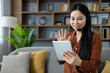 © Liubomir - Asian woman participates in virtual meeting at home using tablet and headset. She is sitting on sofa, waving and smiling, demonstrating technology's role in remote communication.