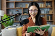 © Liubomir - Asian woman with glasses recording podcast at home, reading book. Engaged in creative broadcasting. Relaxed indoor setting, communicating ideas, sharing knowledge.