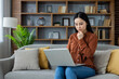 © Liubomir - Asian woman sitting on sofa using laptop, deep in thought, working from home. Surrounded by books and modern decor, conveying focus, contemplation, productivity, home office setting.