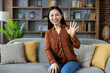 © Liubomir - Asian woman smiling and waving while seated on sofa in modern home. She is dressed casually and appears happy. Room features include stylish shelves and comfortable furnishings.