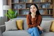 © Liubomir - Asian woman relaxing at home, sitting on sofa with thoughtful expression. Surrounded by books, cushions, and plants, conveying calm and contemplation in a stylish living room setting.