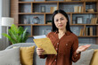 © Liubomir - Asian woman with perplexed look holds envelope, expressing confusion over its contents. Scene captures emotion and curiosity in a cozy home environment, highlighting surprise.