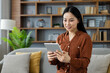 © Liubomir - Asian woman using tablet at home. She is sitting on sofa, smiling, enjoying leisure time with technology. Casual wear enhances cozy atmosphere. Tablet and modern living space prominent.