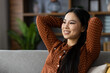 © Liubomir - Asian woman in polka dot blouse enjoying relaxation at home, smiling as she leans back on sofa. Her peaceful expression conveys happiness and contentment in cozy setting.