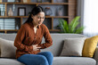© Liubomir - Asian woman on sofa holding stomach in discomfort, expressing pain. Casual brown shirt complements serene home background. Natural light creates a calming atmosphere despite discomfort.