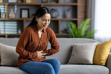 Asian woman on sofa holding stomach in discomfort, expressing pain. Casual brown shirt complements serene home background. Natural light creates a calming atmosphere despite discomfort.