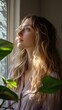 © vadosloginov - Thoughtful young woman gazing out a window surrounded by lush green plants in warm morning light