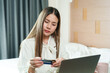 © Ekkasit A Siam - Asian female office worker using credit card for online shopping at home, sitting in cozy bedroom with laptop, highlighting convenience, modern technology, and relaxed lifestyle.