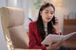 © Tj - Asian businesswoman wearing a stylish red jacket, talking on a mobile phone while reading notes, sitting comfortably in an armchair within a modern office environment