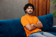 © ViDi Studio - Side view young frustrated sad confused Indian man wears orange casual clothes sits on blue sofa couch stay at home hotel flat rest relax spend free spare time in living room indoor. Lounge concept.