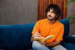 © ViDi Studio - Young Indian man wear orange casual clothes sits on blue sofa couch write down memories in notebook diary stay at home hotel flat rest relax spend free spare time in living room indoor Lounge concept