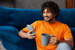 © ViDi Studio - Young Indian man wear orange casual clothes sit near blue sofa couch hold use mobile cell phone drink tea stay at home hotel flat rest relax spend free spare time in living room indoor Lounge concept