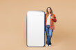 © ViDi Studio - Full body young woman she wears orange shirt casual clothes big huge blank screen mobile cell phone smartphone with workspace copy space mockup area show thumb up isolated on plain beige background.