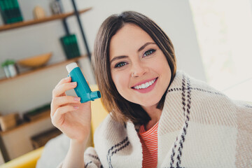 Wall Mural - Young woman smiling indoors holding inhaler, feeling relaxed and comfortable in cozy living room setting