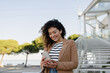 © mary_markevich - pretty curly smiling woman walking in city street in stylish jacket, using smartphone
