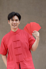 Portrait of happy smiling Asian man in red traditional Chinese dress holding red envelopes in front of cement wall. Handsome Chinese male showing Ang Pao to celebrate Lunar Chinese New Year.