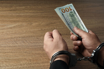 Wall Mural - Corruption. Man in handcuffs with dollar banknotes at wooden table, closeup. Space for text