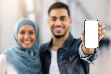 Wall Mural - Beat App. Cheerful Islamic Couple Showing Smartphone With Blank Black Screen At Camera While Standing In Airport Terminal, Happy Muslim Spouses Recommending New Mobile Application, Mockup Image