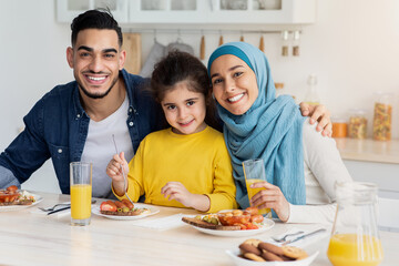 Wall Mural - Portrait Of Happy Muslim Family Of Three Posing At Camera While Eating Breakfast In Kitchen Together, Cheerful Middle Eastern Parents And Their Little Daughter Sitting At Table Enjoying Tasty Food