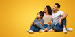 © Prostock-studio - Happy Young Arab Family Of Three Holding Digital Tablet And Looking Aside With Interest, Middle Eastern Parents And Little Daughter Sitting On Floor Over Yellow Background, Panorama With Copy Space