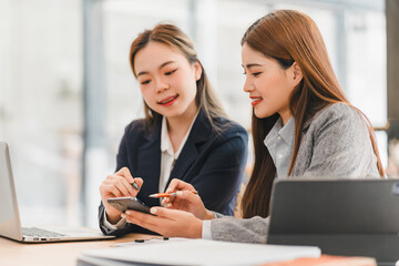 Wall Mural - Two businesswomen discussing work while using smartphone and laptop in modern office setting, showcasing teamwork and collaboration