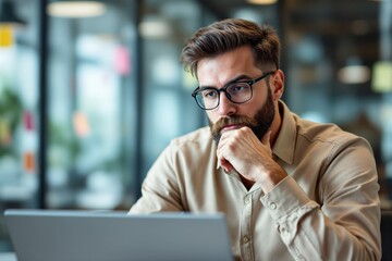 Poster - Thoughtful man with glasses working on a laptop in modern office environment.