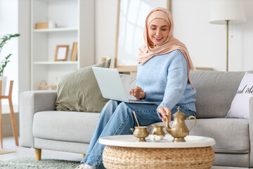 Poster - Young Muslim woman with laptop and Turkish tea sitting on sofa at home