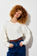 Wall Mural - Smiling woman with curly hair in a cozy white sweater and high waisted blue jeans poses confidently against a light background Her joyful expression brings warmth to the image