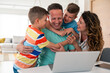 © Dorde - Caucasian professional in turquoise casual shirt works on laptop at home while family members share affectionate moments in bright living space.