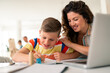 © Dorde - Caucasian mother with dark curly hair assists young boy in colorful shirt with schoolwork at bright home study space with laptop nearby.
