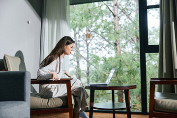 Canvas Print - Young woman working on a laptop in a bright room, displaying emotions of concentration and determination in a natural setting with greenery