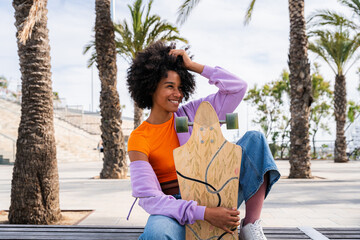 Wall Mural - Beautiful young black woman with curly afro hair portrait outdoors - Modern cool female wearing stylish urban clothing strolling in the city with longboard