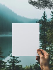 Poster - Blank paper held in hand against a tranquil lake and forest backdrop during a misty morning. Generative AI