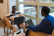 © Studio Marmellata - Two young black men laughing while working on laptops in a cozy cafe, seated on wicker chairs near large windows, creating a lively and engaging environment filled with natural light