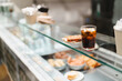 © Studio Marmellata - A glass counter in a cafe displays a slice of cake and a glass of iced drink alongside other baked goods, offering a glimpse of the appealing and carefully curated menu items in a modern setting.
