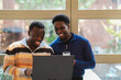 © Studio Marmellata - Two black friends in a cafe look at a laptop together near large windows with natural light and greenery outside, enjoying a collaborative and engaging moment in a modern and cozy environment.