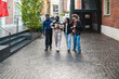 © Studio Marmellata - A group of four young adults walks together on a cobblestone street, enjoying a friendly conversation while wearing casual outfits, with urban buildings and greenery in the background.