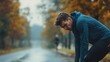 © vefimov - A male athlete in a blue jacket and black shorts is crouched over on the side of a road, leaning against a fence. The scene captures a moment of pause or recovery during a running training session