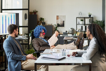 Poster - Clown boss keeping legs on desk and looking through financial papers while sitting in front of two young economists
