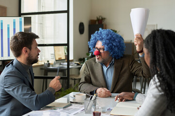 Poster - Angry mature boss in formalwear, clown wig and nose expressing irritation while sitting in front of two subordinates and speaking to one of them
