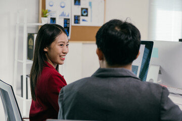 Poster - Two asian businesspeople are working together in a modern office, with a woman turning to smile at her male colleague who is concentrating on his computer screen