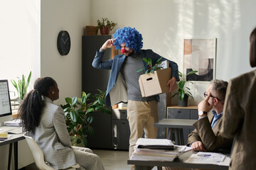 Poster - Young funny male office worker in clown wig holding box with supplies and looking at female colleague while leaving