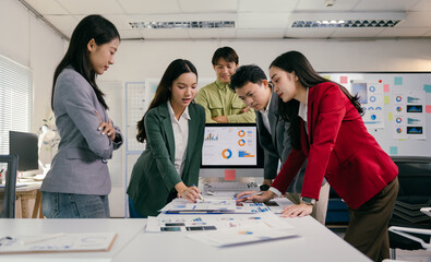 Wall Mural - Diverse group of young asian professionals analyzing graphs and reports, collaborating on a project, using computer and printed documents in a modern office environment