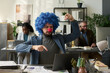 © pressmaster - Young male analyst in formalwear and clown wig sitting by workplace in front of laptop and pressing keys during network
