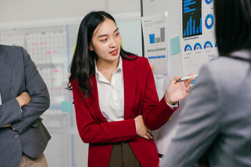 Aufkleber - Young asian businesswoman wearing red blazer pointing at charts on whiteboard while leading a meeting with colleagues, discussing marketing data and company strategy
