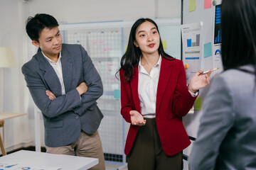 Poster - Asian businesswoman in a red blazer leads a meeting. Presenting charts and data on a whiteboard to her colleagues in a modern office setting. Fostering collaboration and strategic decision-making