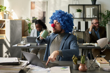Poster - Young businessman in clown wig looking at mug with coffee and pressing key of laptop keypad while having break in office