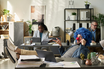 Poster - Young male employee in blue curly wig and red nose speaking on mobile phone while relaxing by workplace with laptop and papers