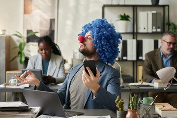 Poster - Young clown employee with smartphone in hand watching online yoga course and relaxing by workplace in pose of lotus